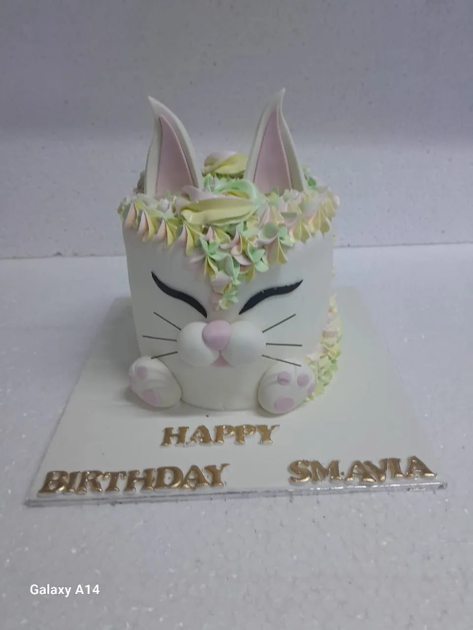 3D white cat face birthday cake with pink fondant paws, ears, and pastel piped cream crown on a white cake board by Sweet Cake Studio.
