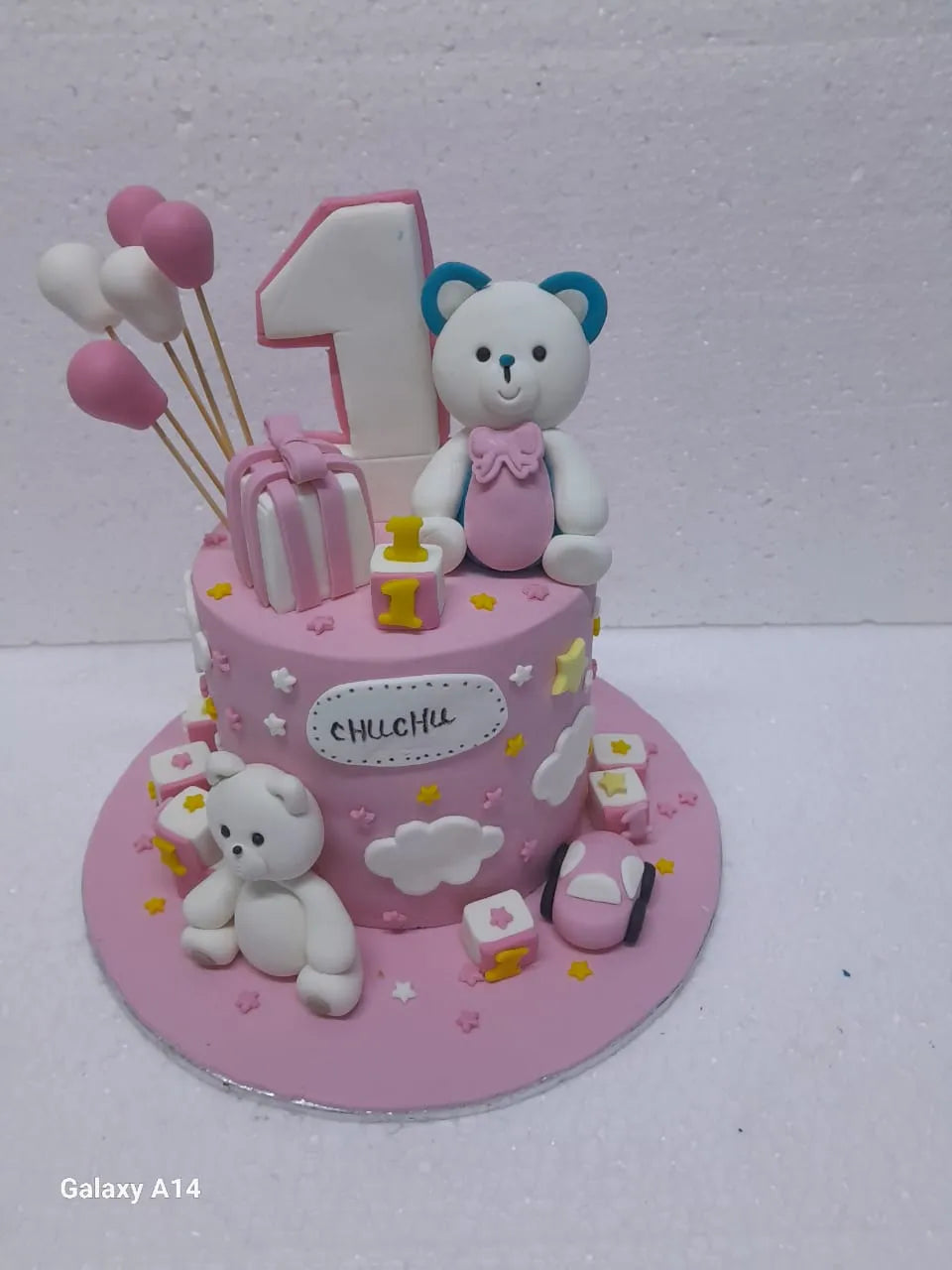 A beautiful light pink fondant 1st birthday cake decorated with handcrafted white and pink teddy bears, fondant alphabet blocks, miniature balloons, and clouds by Sweet Cake Studio.