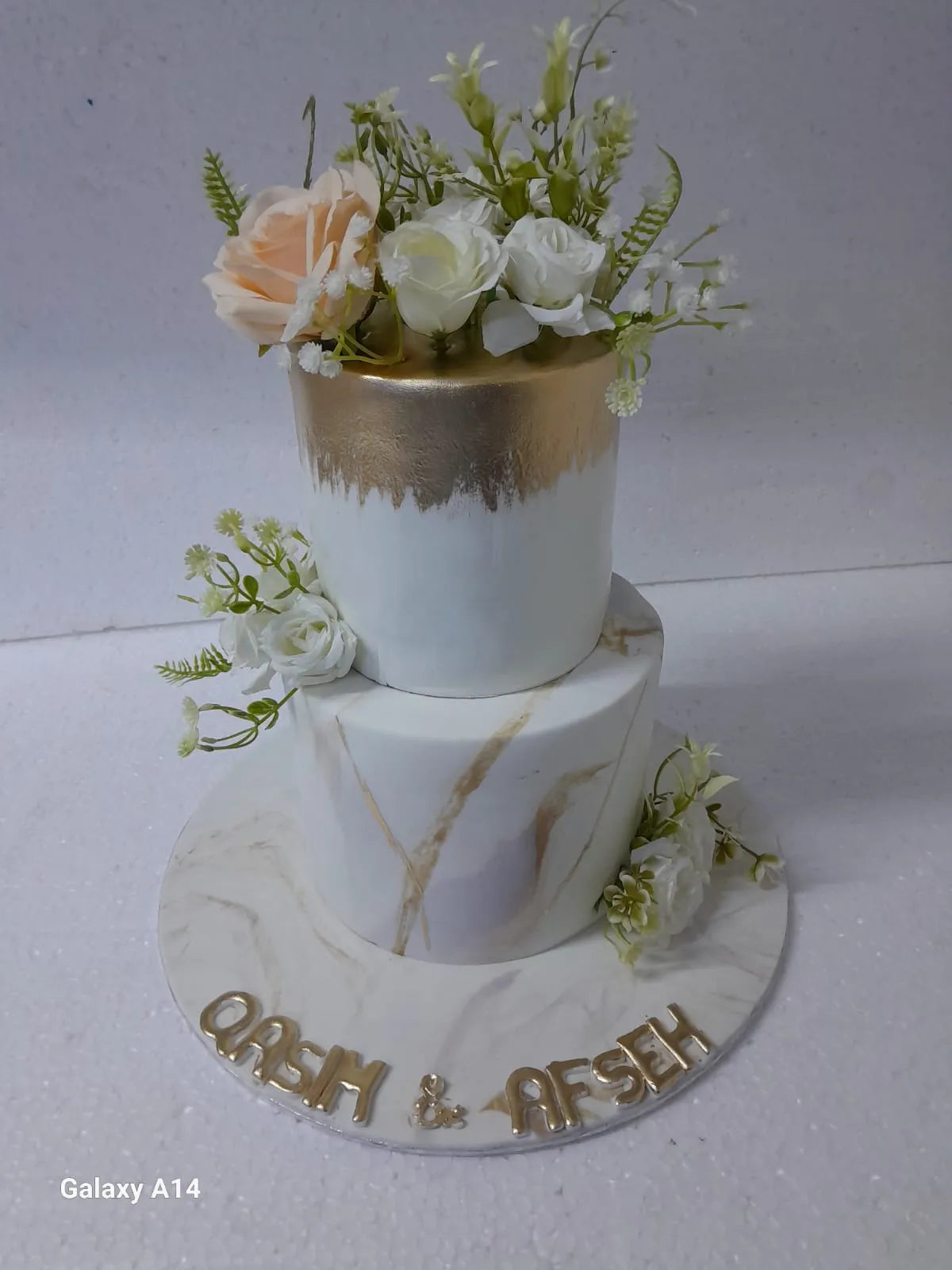 A sophisticated 2-tier white fondant cake with a gold-painted metallic top edge, realistic gold marble veins on the bottom tier, and delicate white and peach roses by Sweet Cake Studio.