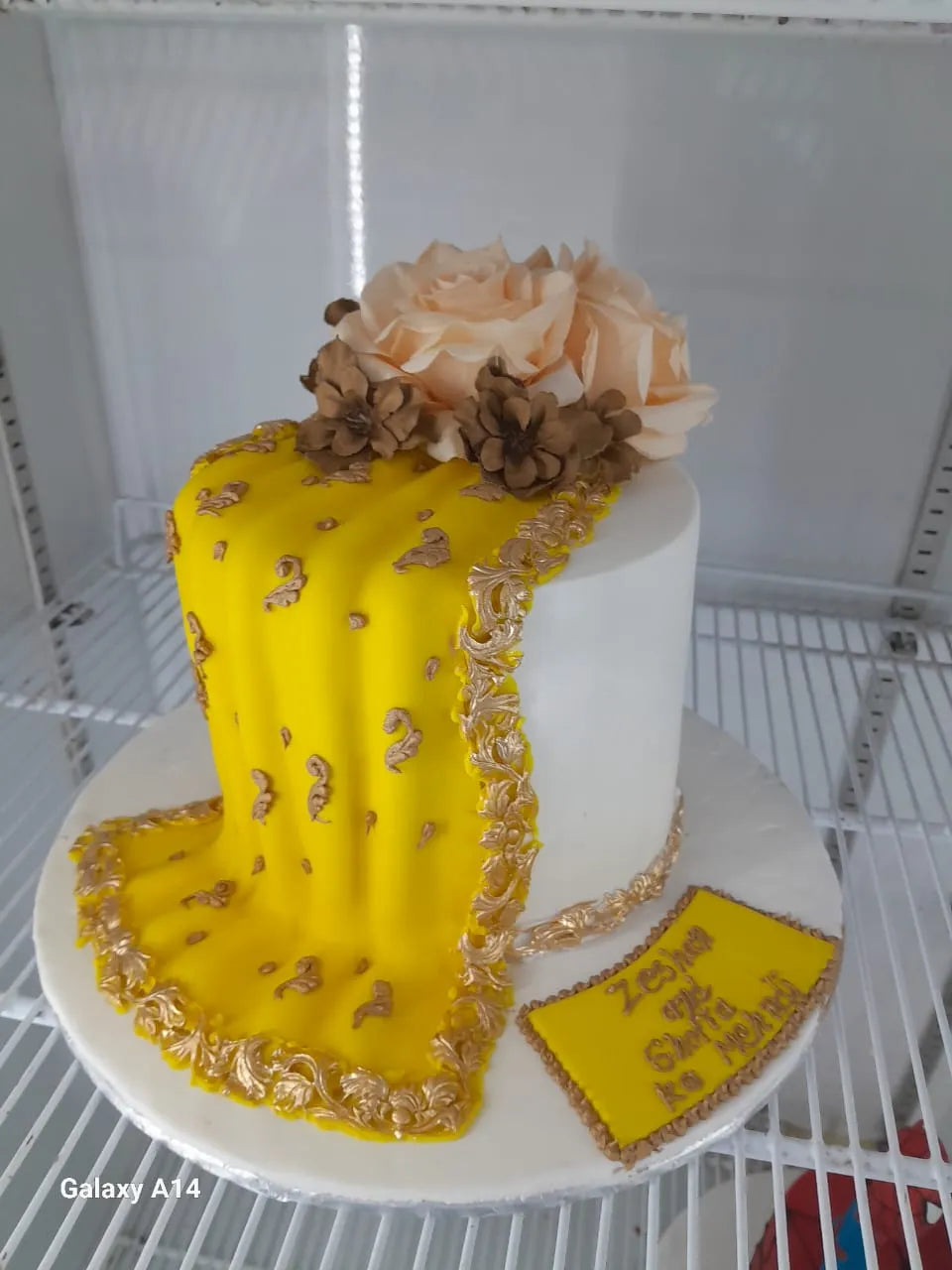 A beautiful white celebration cake featuring an elegant bright yellow fondant bridal drape designed like a traditional dupatta with intricate hand-painted golden borders and motifs, topped with large peach roses by Sweet Cake Studio.