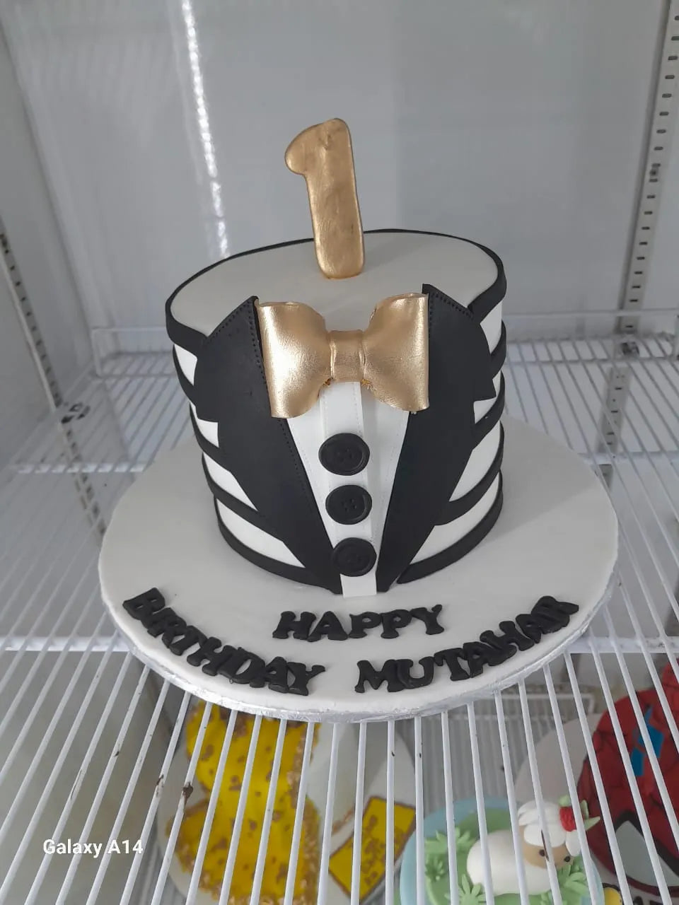 Cake designed like a tuxedo with a bow tie and number 1 on top, placed on a white surface.