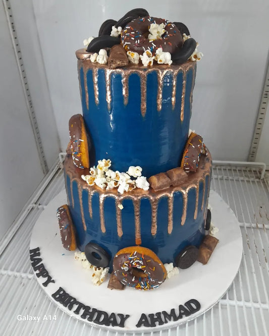 Two-tiered birthday cake with blue icing, chocolate elements, and popcorn on a white stand.