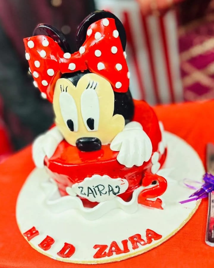 Minnie Mouse-themed birthday cake with red bow and 'Zaira' text on a red surface.
