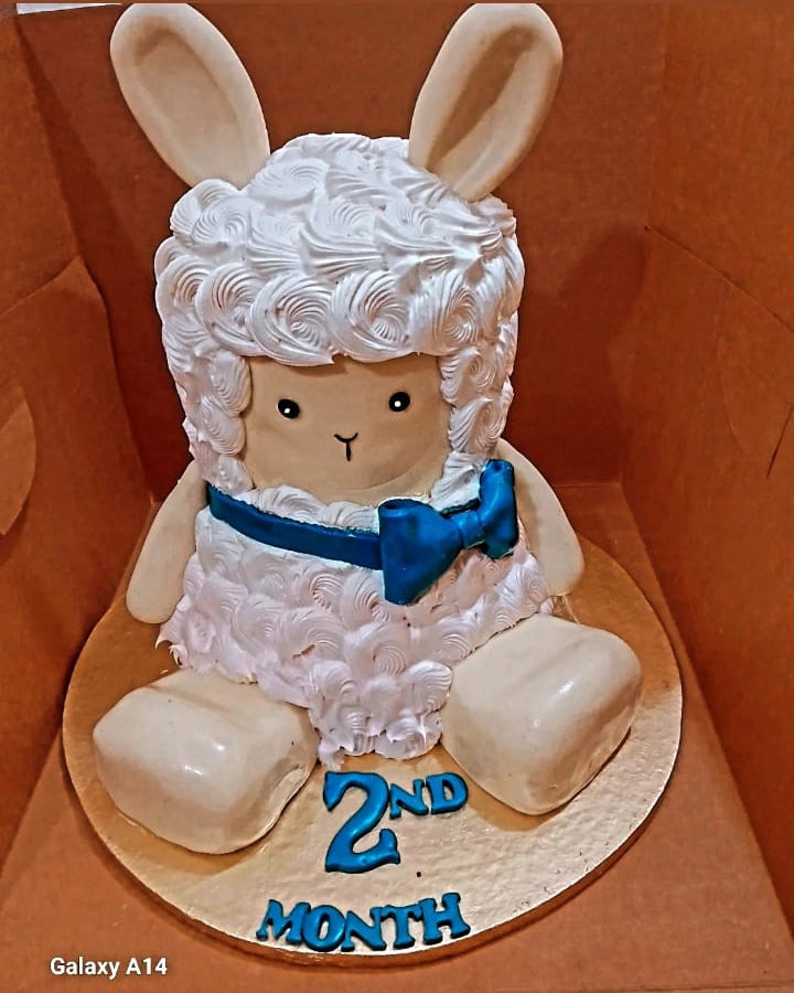 Sheep-themed cake with white fur, blue bow tie, and '2nd Month' text on a brown background